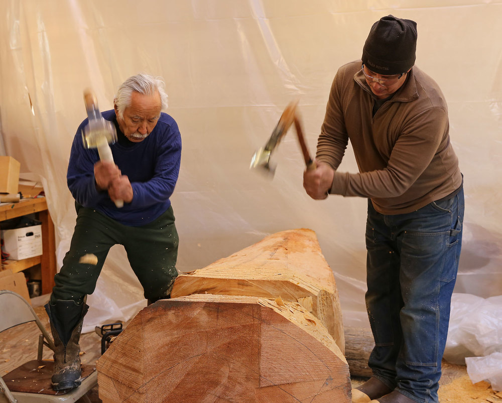 Nathan Jackson and John Hughes using adzes to shape a cedar log into a totemic sculpture.